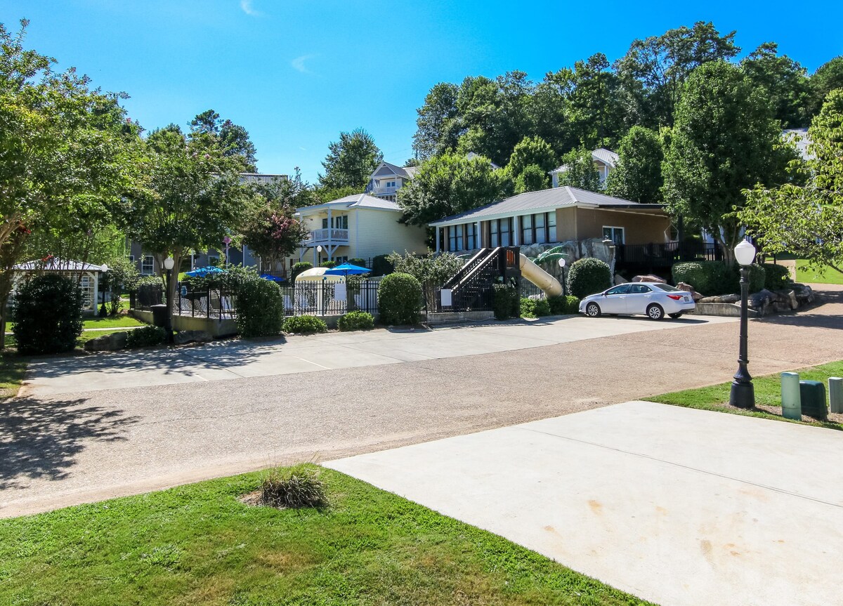 A spacious area features well-maintained landscaping with trees and shrubs surrounding a paved driveway. Multiple structures are visible, including a house and recreational facilities. A parking space for vehicles is present, providing convenient access to the property's amenities.