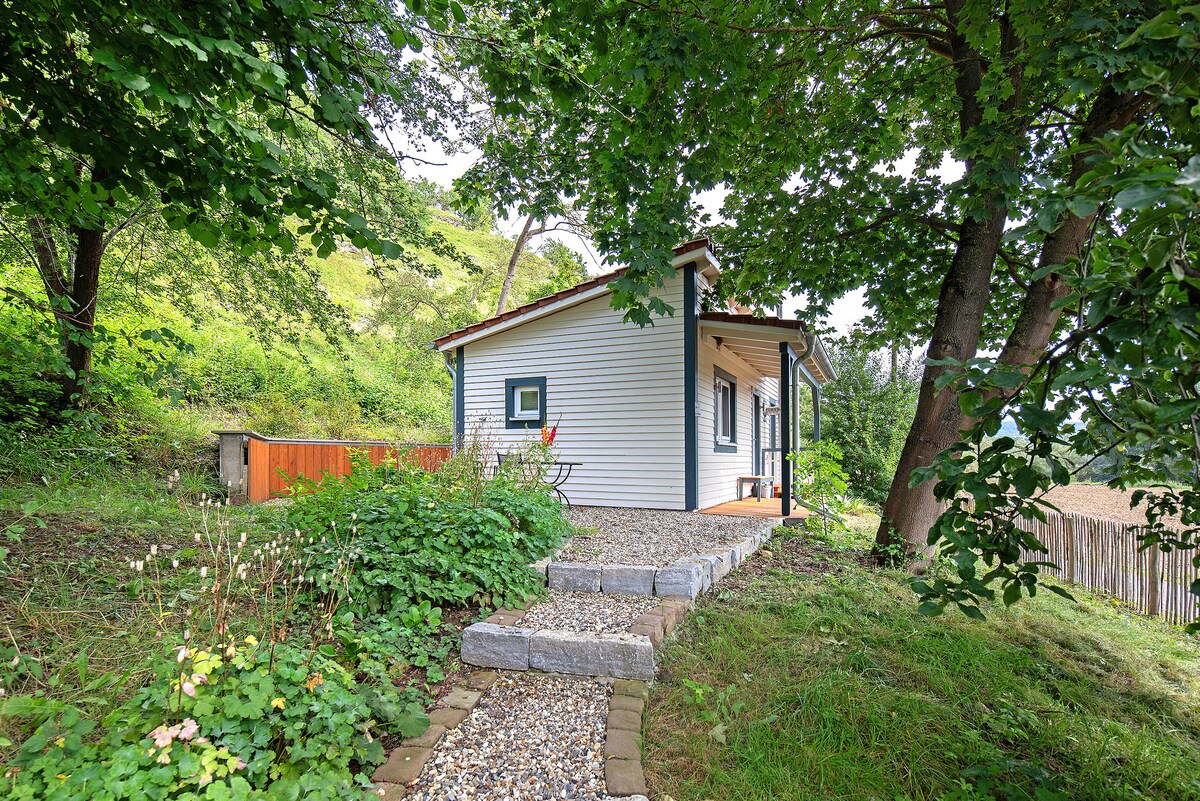 A pathway lined with gravel leads to a charming vacation home nestled among lush greenery. The structure features a light-colored exterior, with large windows that invite natural light. A cozy outdoor seating area is visible beside the home, enhancing the serene environment.