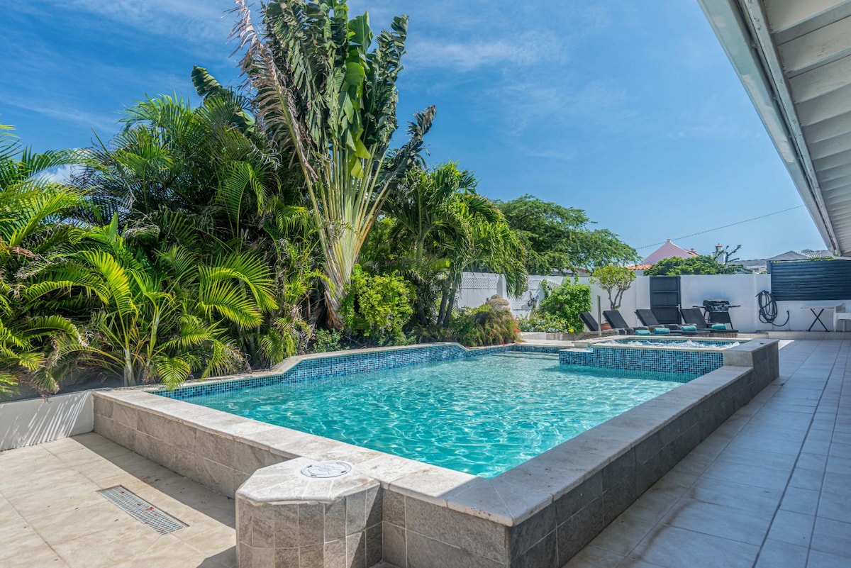 A tranquil outdoor space features a sparkling pool surrounded by lush tropical plants. An adjacent whirlpool tub adds to the relaxation options, while sun loungers are positioned nearby, inviting guests to enjoy the serene atmosphere.
