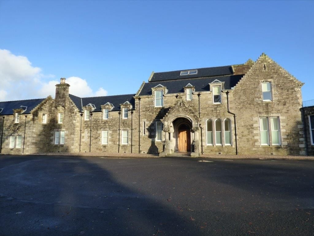 The stone exterior of Lomond Castle features intricate architectural details and multiple windows, reflecting a blend of historical charm and modern design. The entryway is accentuated by a large wooden door, flanked by arched windows, against a clear blue sky.