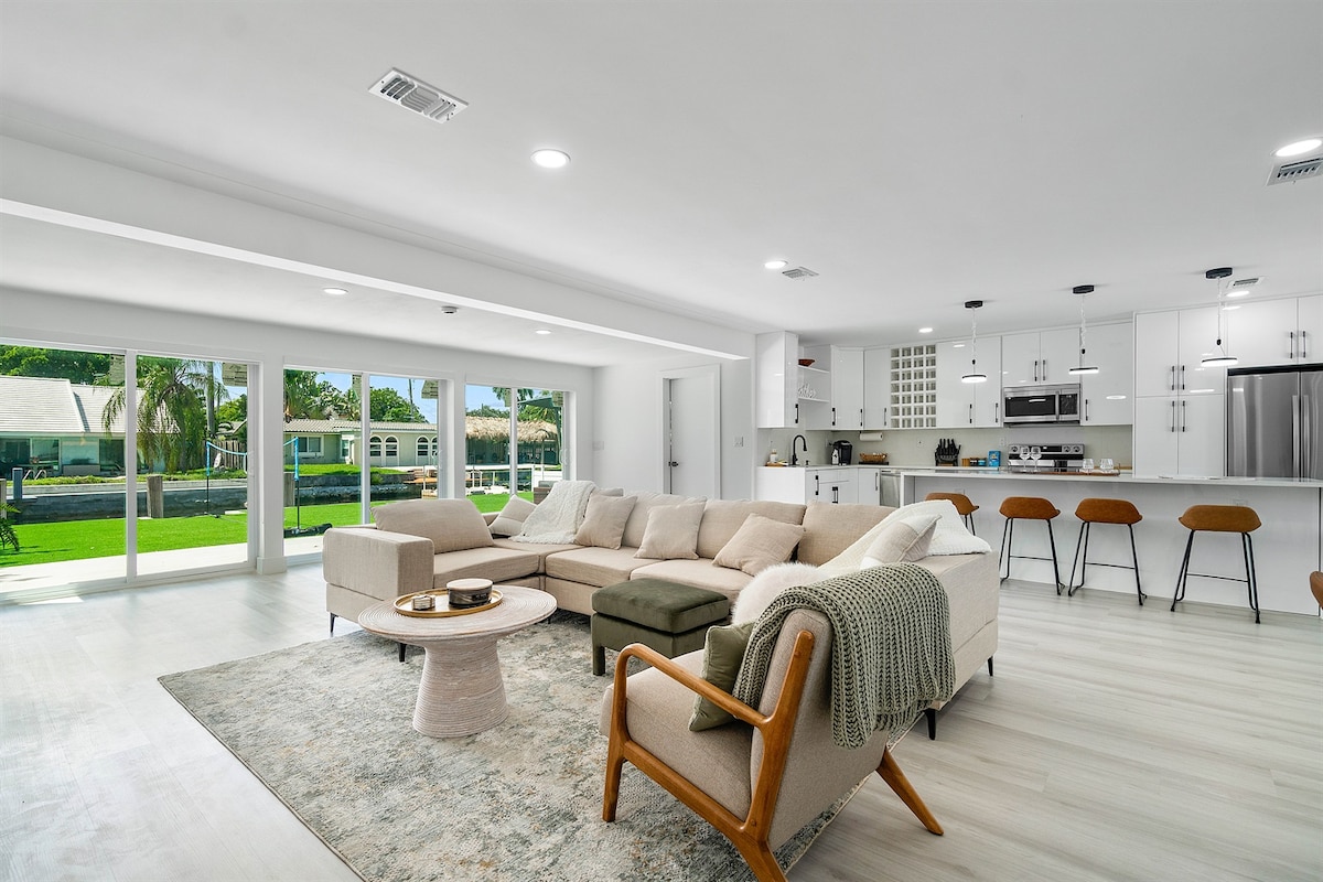 The spacious living area features a sectional sofa in neutral tones, complemented by a round coffee table. Adjacent to the living space, a modern kitchen with sleek cabinetry and bar stools can be seen. Two large sliding glass doors flood the room with natural light and provide a view of the backyard.