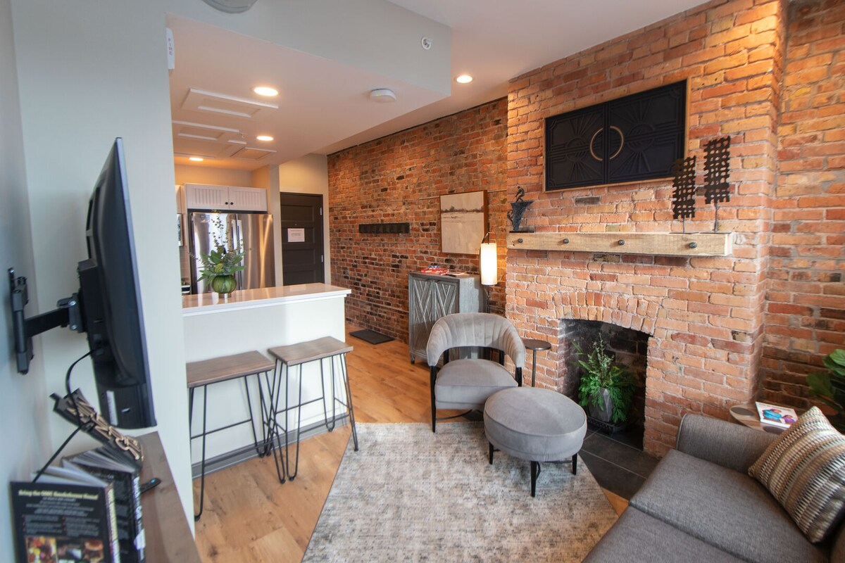 The living area features exposed brick walls and elegant decor, creating a warm ambiance. A comfortable gray armchair and a cozy sectional sofa are positioned near a sleek entertainment unit. A stylish coffee table sits atop a textured area rug, complementing the chic design.