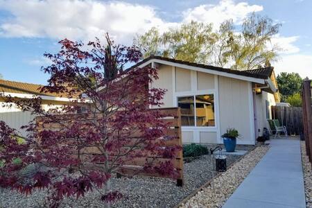 A single-story guest suite is nestled in a tranquil setting, featuring a wooden fence and a neat gravel landscape. A small tree with reddish leaves adds a touch of color, while a potted plant sits near the entrance. Comfortable seating is visible beside the doorway.