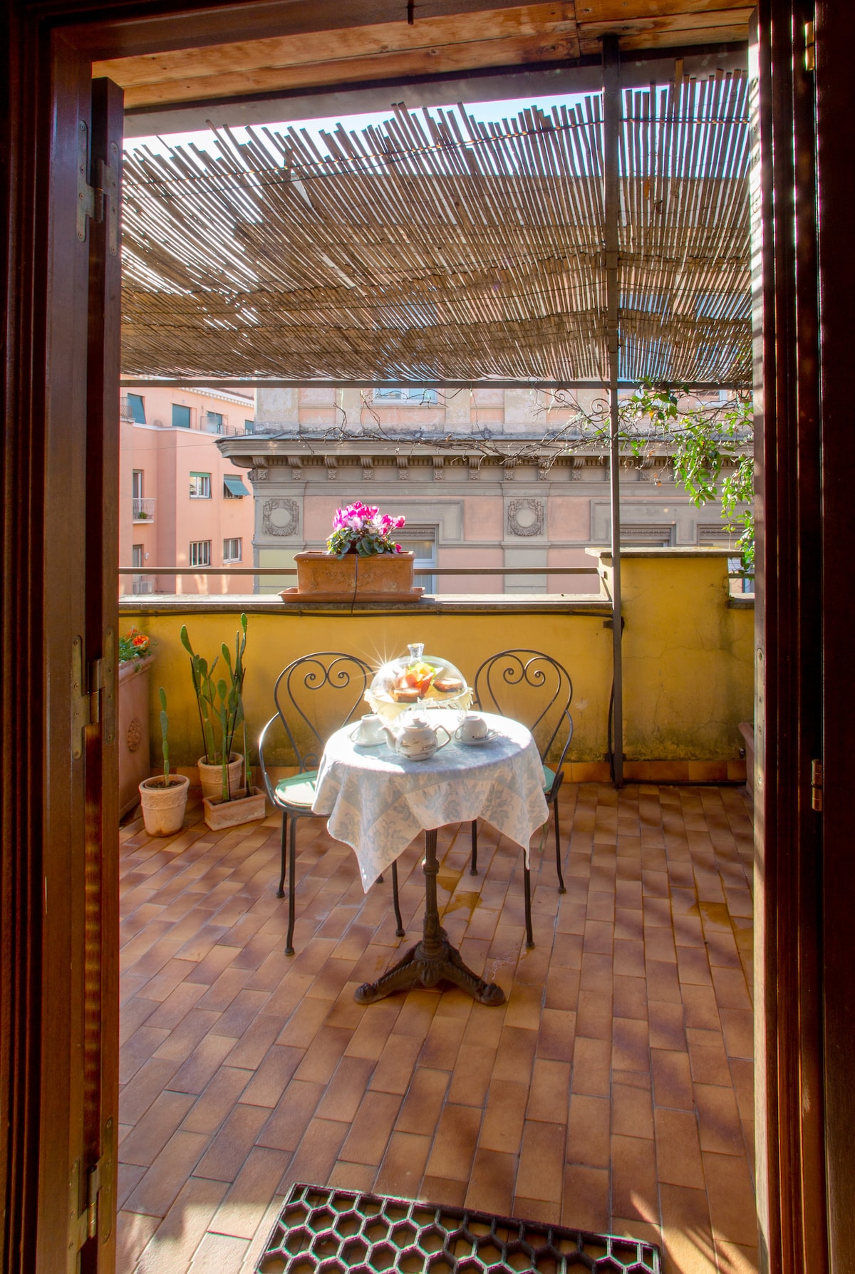 A terrace is viewed through open wooden doors, featuring a small round table with two chairs. The table is set with a tea service and pastries, surrounded by potted plants. A bamboo shade offers partial coverage, creating a cozy outdoor space.