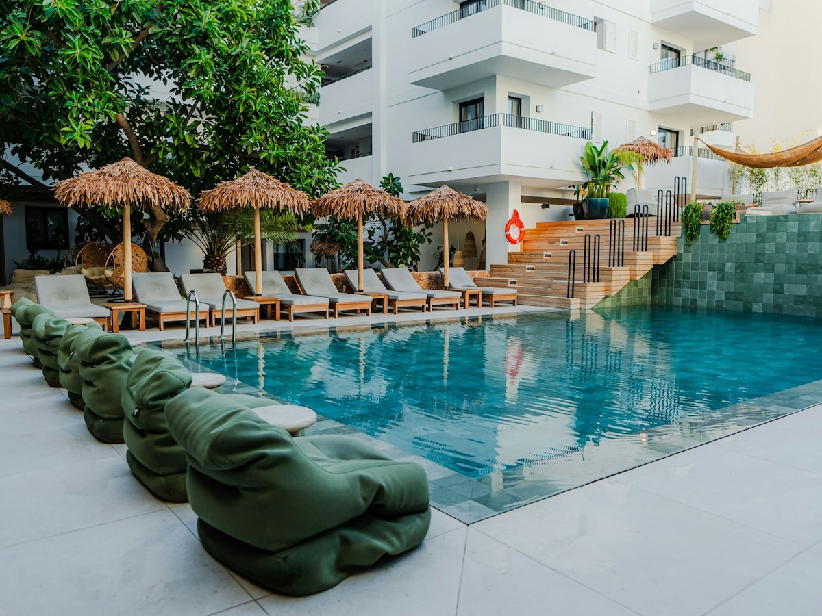 An outdoor pool area features a calm, clear water surface and is framed by green bean bag seating. Several sun loungers with thatched umbrellas provide shade. Stairs lead to a wooden deck, surrounded by tropical greenery and an inviting hammock.