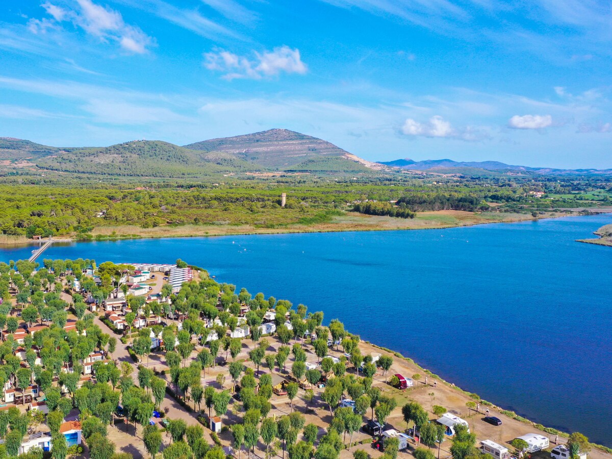 An aerial view captures the expansive Camping Village Laguna Blu, nestled among lush greenery. The serene blue waters of a lake reflect the surrounding mountains, while the property layout showcases numerous mobile homes within the tree-filled resort, providing a tranquil environment.