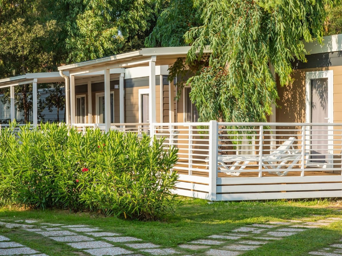 The exterior of a mobile home is set amid lush greenery, featuring a shaded terrace with lounge chairs. A pathway of stone tiles leads to the home, framed by vibrant shrubs and trees that provide a natural ambiance.