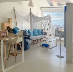A living area features a cozy seating arrangement with a light-colored sofa adorned with various cushions. A sheer fabric drapes overhead, contributing a soft ambiance. Large glass doors provide a view of the ocean, while a simple glass coffee table and stylish bar stools enhance the space.