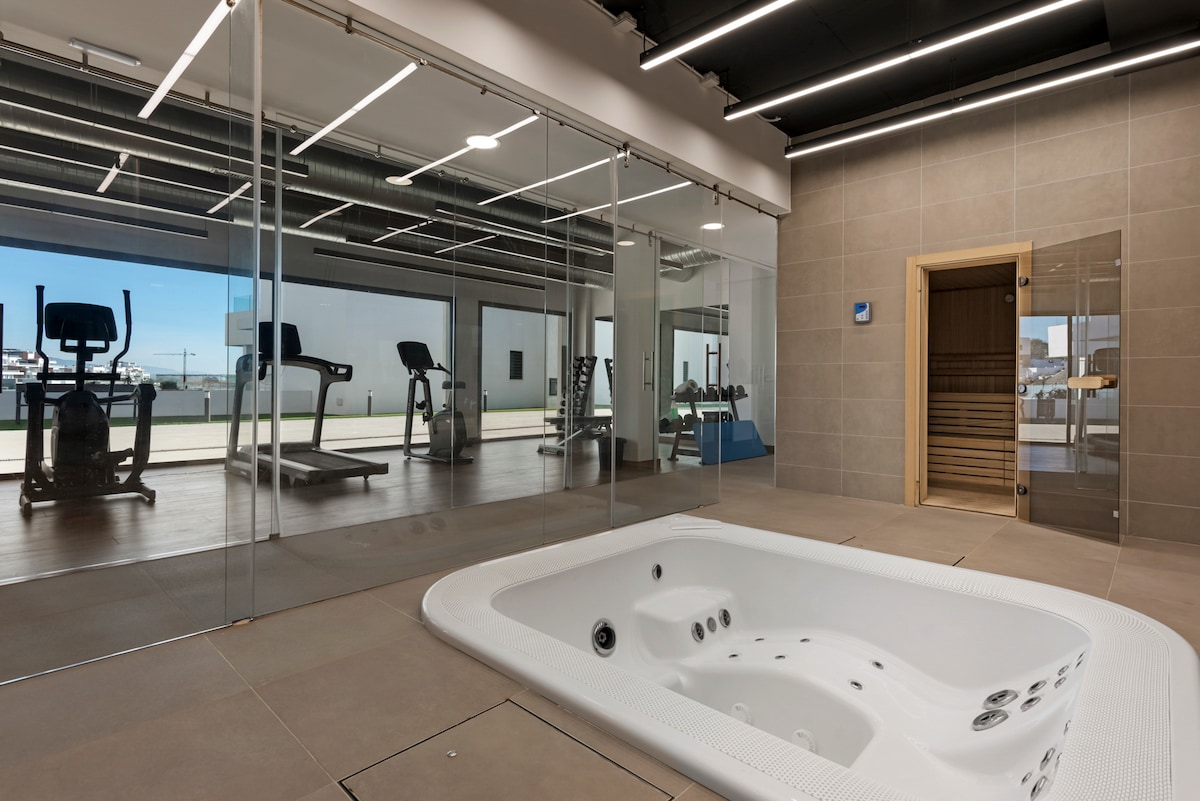 A modern wellness area features a jacuzzi alongside large glass windows. An adjacent fitness space is equipped with exercise machines, while a sauna is tucked away in the corner. Natural light enhances the open, clean lines of the design.