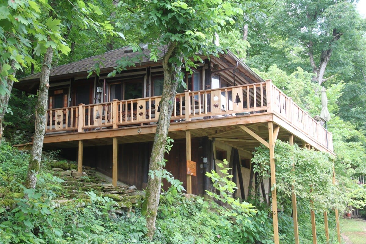 The cabin is elevated on a wooden deck surrounded by lush greenery. The structure features large windows and a welcoming entrance, while the deck is furnished with chairs and tables, inviting relaxation and outdoor enjoyment.