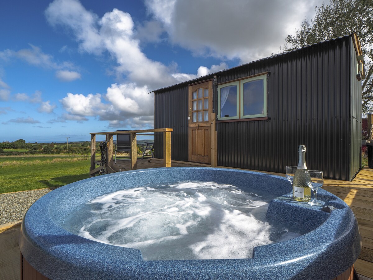 A private hot tub filled with bubbling water is positioned on a wooden deck, surrounded by open green space. The well-maintained exterior of a shepherd's hut is visible in the background, with a wooden door and large window framed by a blue sky and scattered clouds.