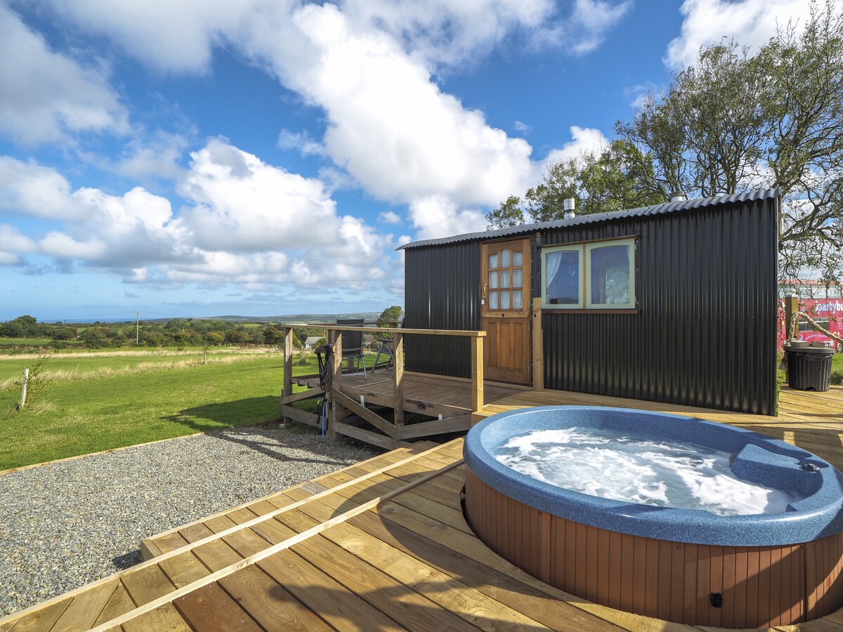 A shepherd's hut with a dark exterior and wooden deck is surrounded by lush greenery. A circular hot tub is situated on the deck, inviting relaxation. The sky above features a mix of clouds and blue, with views extending towards distant hills.