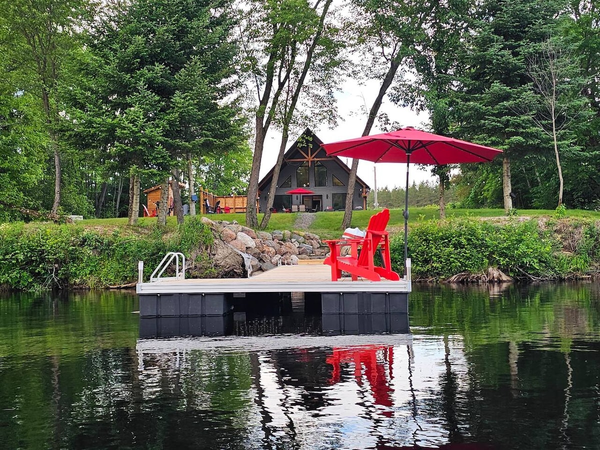 A small dock extends into calm water, featuring two red Adirondack chairs and a matching umbrella for shade. Lush greenery surrounds the dock, reflecting on the water's surface. In the background, the chalet's angled roof and outdoor seating space are visible, framed by trees.