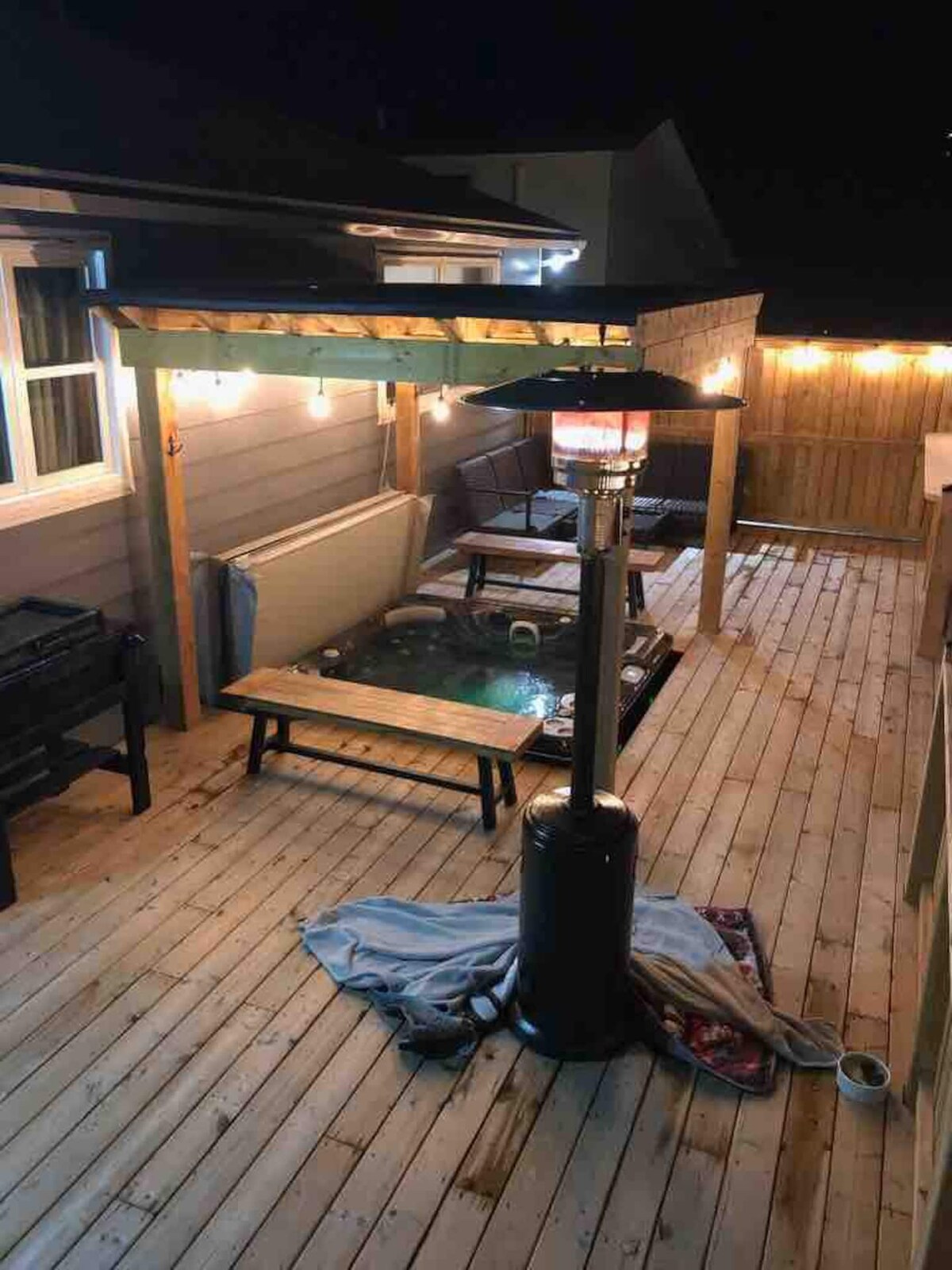 An outdoor space is featured with a hot tub, surrounded by wooden decking. A propane fire pit stands nearby, and string lights create a soft glow. Several seating areas with cushions invite relaxation, while a towel is draped over the edge of the deck.