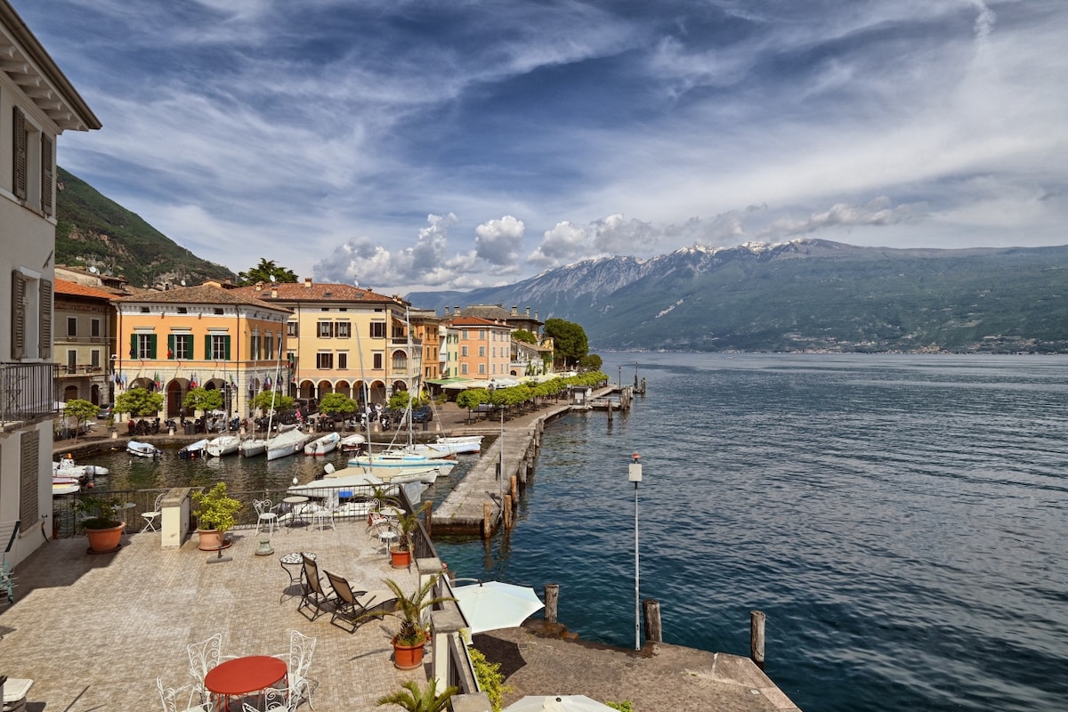 A serene view of the waterfront in Gargnano features colorful buildings lining the port. Calm waters reflect the surrounding mountains and cloudy blue sky. Several boats are moored along the pier, while shaded seating areas are positioned along the walkway, inviting relaxation by the lake.