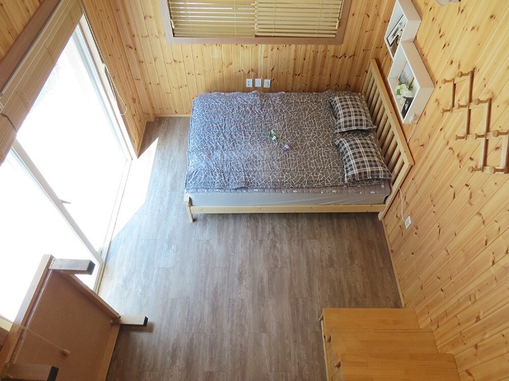 The room features a spacious bed adorned with a subtle patterned blanket and two neatly arranged pillows. Natural light pours in through large sliding glass doors, highlighting the warm wooden interior. A simple wooden dresser and a clothes rack are positioned nearby.