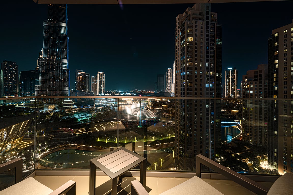 A balcony terrace is shown with modern outdoor seating, offering views of the illuminated skyline of Downtown Dubai. The Burj Khalifa is prominently visible, along with sparkling lights from surrounding buildings and the vibrant cityscape reflecting on water features below.