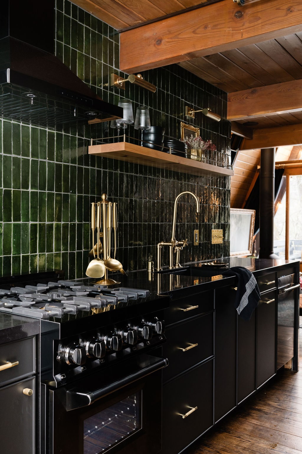 A modern kitchen features dark cabinetry complemented by a green tiled backsplash. A gas range with multiple burners is prominently displayed, alongside elegant gold fixtures. Open shelving showcases glassware, while a minimalist design reflects a blend of functionality and style within the cabin.