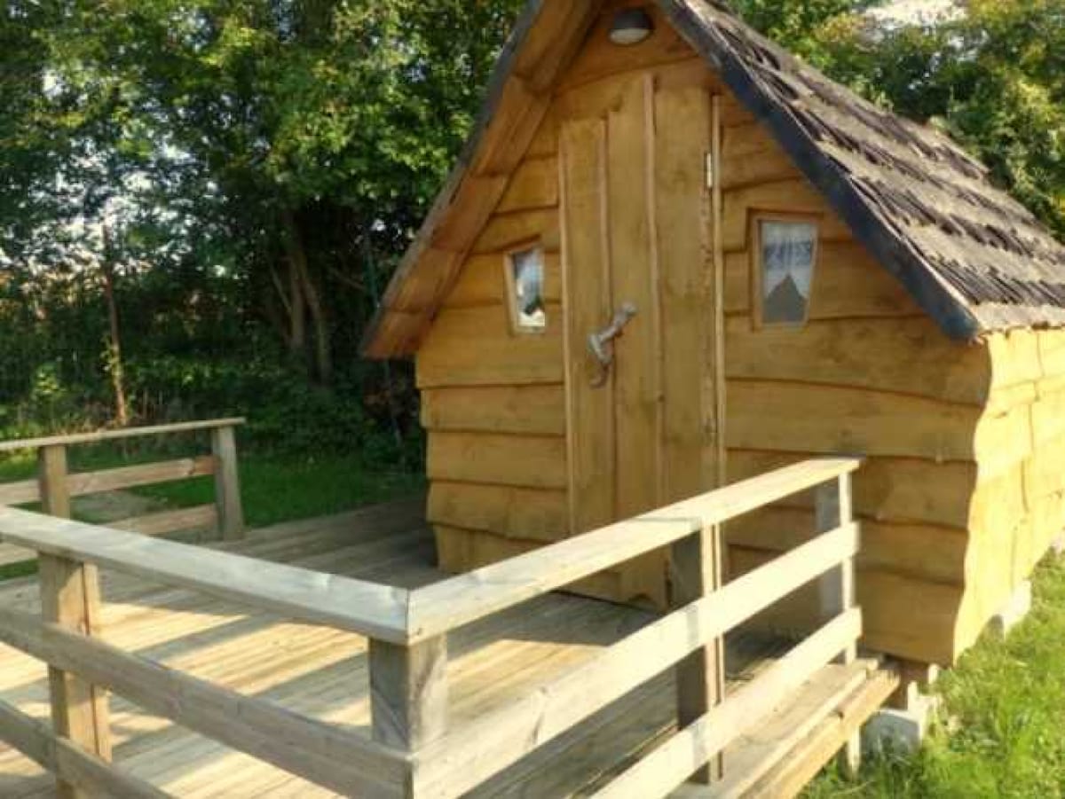 A charming wooden cabin with a sloping roof is set on a wooden deck, surrounded by greenery. The entrance features a sturdy door, flanked by small windows, allowing natural light to illuminate the interior space.
