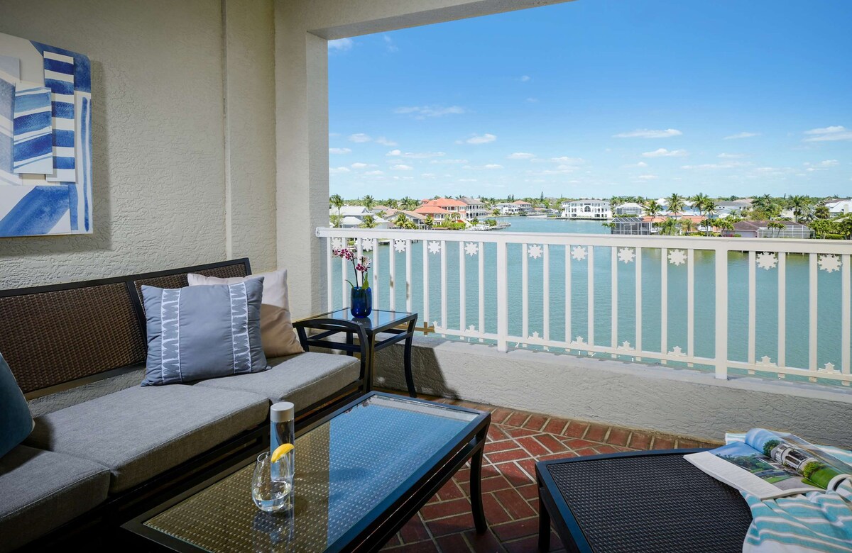 A private balcony features comfortable seating, a glass-topped table, and decorative cushions, offering views of the water and surrounding properties. Potted flowers add a touch of color, while a pitcher and glass of beverage sit on the table, enhancing the relaxing atmosphere.