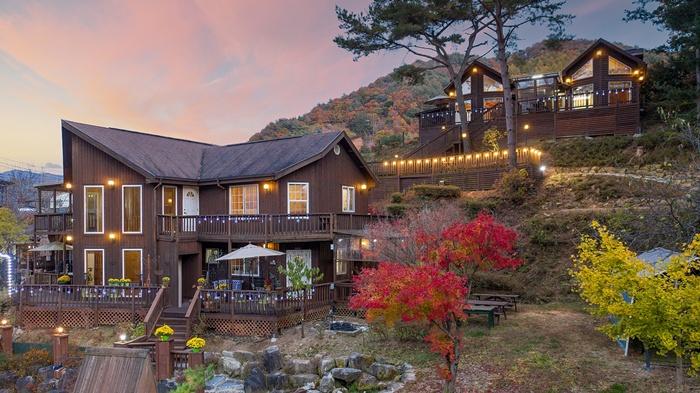 A two-story wooden house is set against a backdrop of hills, with nearby separate structures visible. Outdoor seating areas are present, surrounded by seasonal trees in hues of yellow and red. Soft lighting outlines the buildings, creating a welcoming evening ambiance.