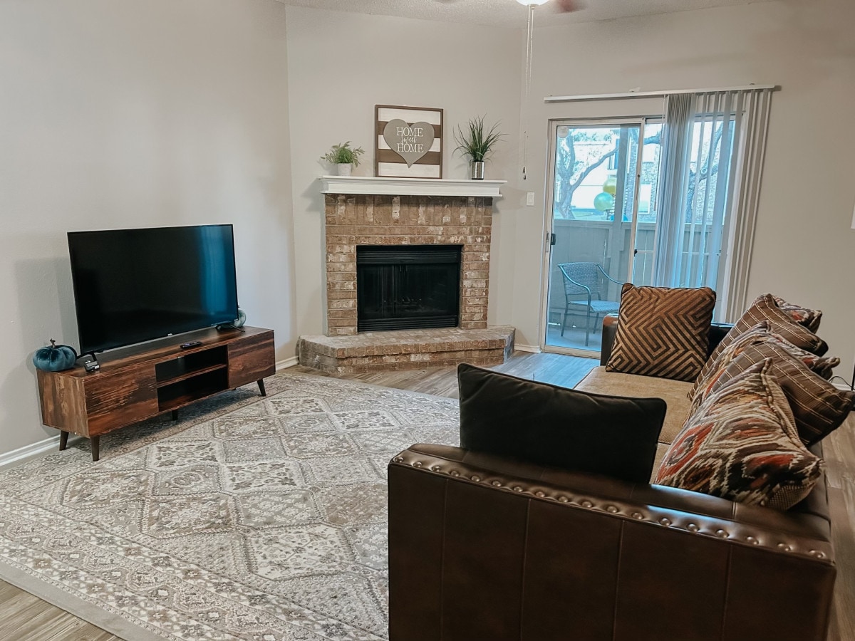 The living room features a comfortable couch adorned with patterned cushions, a wooden entertainment center with a large flat-screen TV, and a classic brick fireplace. Natural light is welcomed in through sliding glass doors that lead to a small outdoor area, enhancing the cozy ambiance.
