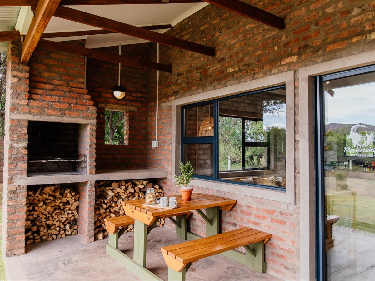 A covered patio area features a built-in braai against a wall of rustic brick. A sturdy wooden table with benches is positioned nearby, and stacked firewood is neatly arranged for convenience. Large windows provide a view of the surrounding landscape.