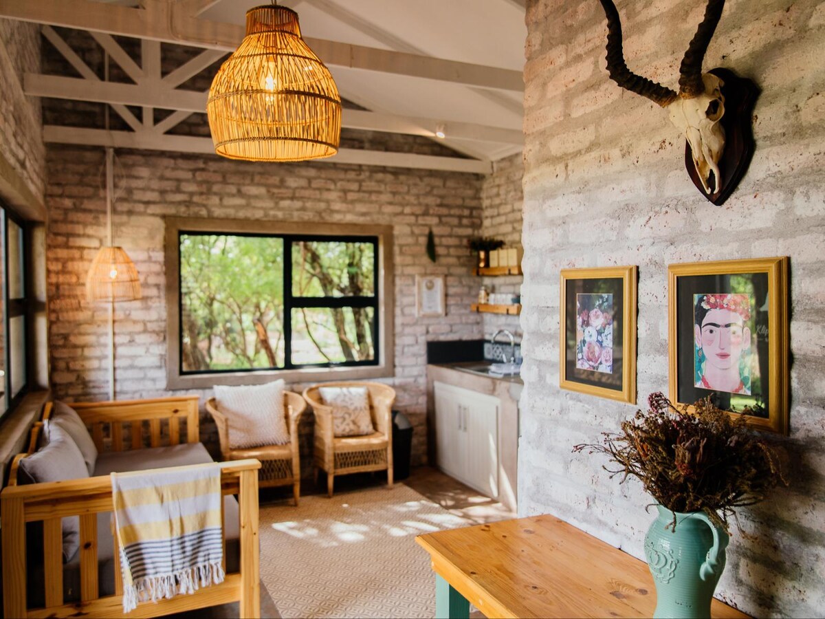 The interior of a cozy chalet is featured, with a light-colored brick wall and wooden beams. A kitchenette is visible alongside a seating area with Malawian wicker chairs. Decorative elements include framed artwork and a mounted antler. Natural light cascades through large windows.
