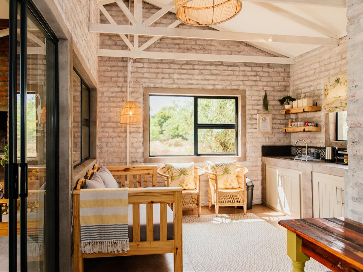 The interior of the chalet features light-toned brick walls and wooden beams. A cozy seating area contains a daybed and two wicker chairs, complemented by a kitchenette with essential amenities. A large window offers views of the surrounding greenery, bathing the space in natural light.