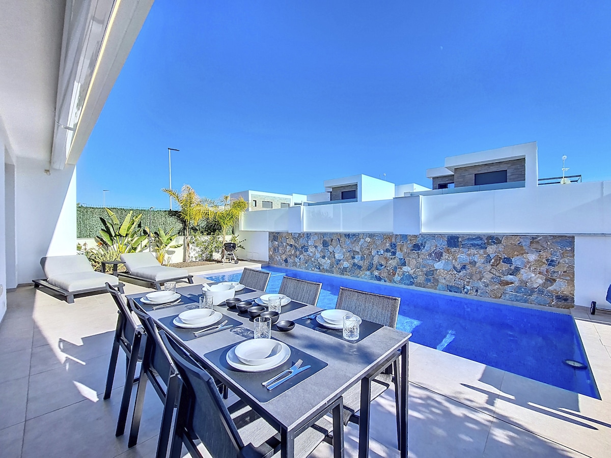 An outdoor dining area is set with a table arranged for six, accompanied by sleek black chairs. A private pool with a stone feature wall is visible in the background, framed by landscaped greenery under a clear blue sky. Sun loungers are positioned nearby.