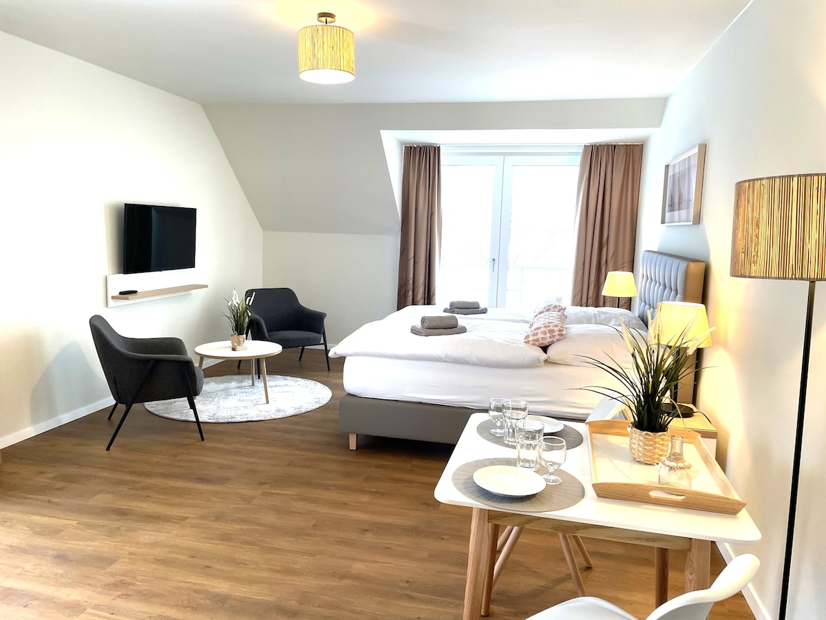A spacious living and sleeping area features a comfortable bed, a lounge chair, and a small coffee table. A modern flat-screen TV is mounted on the wall. Natural light enters through large windows adorned with light curtains, creating a bright and inviting space.