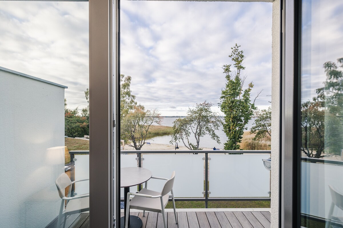 A private balcony features modern outdoor furniture, overlooking a serene landscape with trees and water. The space is framed by sliding glass doors, enhancing views of the natural surroundings and inviting fresh air into the apartment.