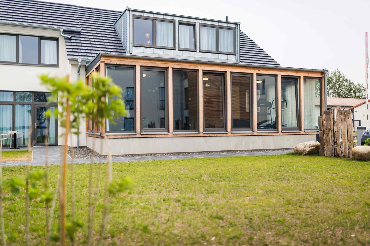A modern vacation house features a large glass-fronted area, allowing natural light to fill the interior. The house is surrounded by a well-maintained lawn, with a small tree and decorative stones visible in the foreground.