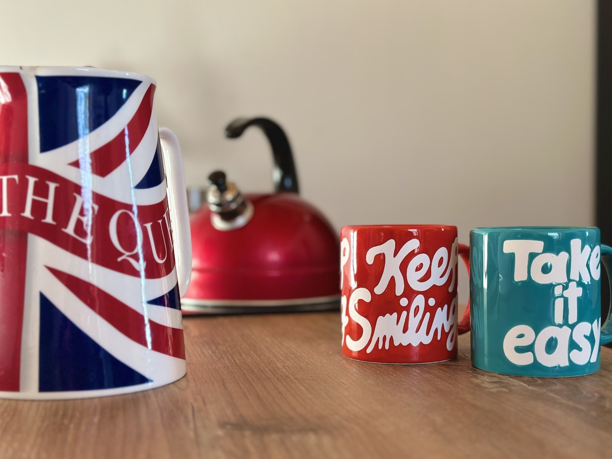 A vibrant kitchen scene is displayed, featuring a large mug adorned with a Union Jack pattern and two smaller mugs with motivational phrases. A red kettle is visible in the background, adding a pop of color against a wooden countertop.