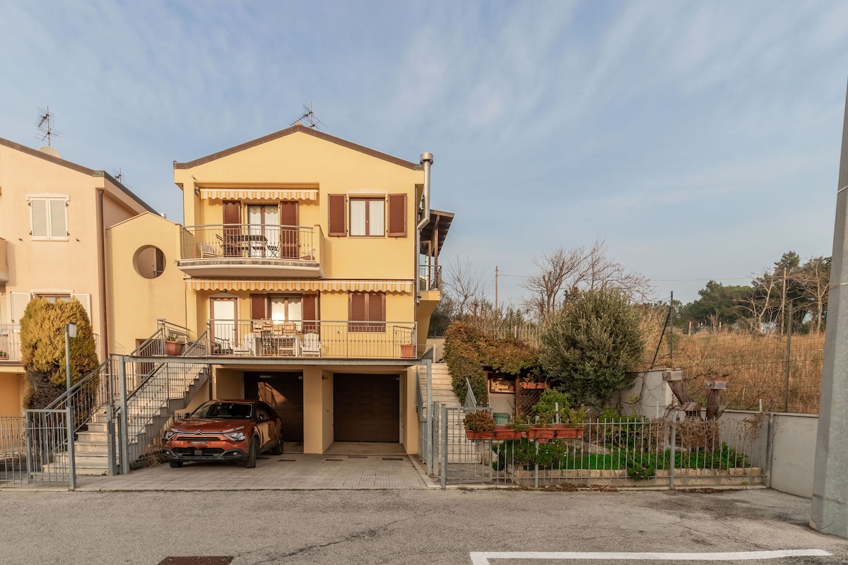 A semi-detached terraced house is presented with light-colored exterior walls. The building features a balcony on the upper floor, along with a covered parking space beneath. Surrounding greenery and a quiet residential area contribute to a calm setting.