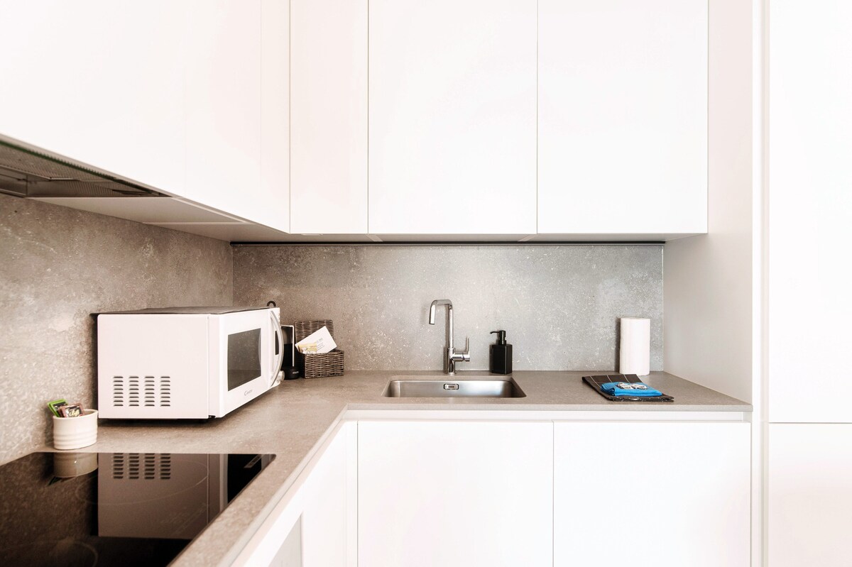 The modern kitchen features a sleek design with white cabinetry and a gray countertop. A microwave, sink, and stovetop are visible, alongside neatly arranged kitchen essentials. A small basket holds miscellaneous items, and a stack of linens is placed on the countertop.