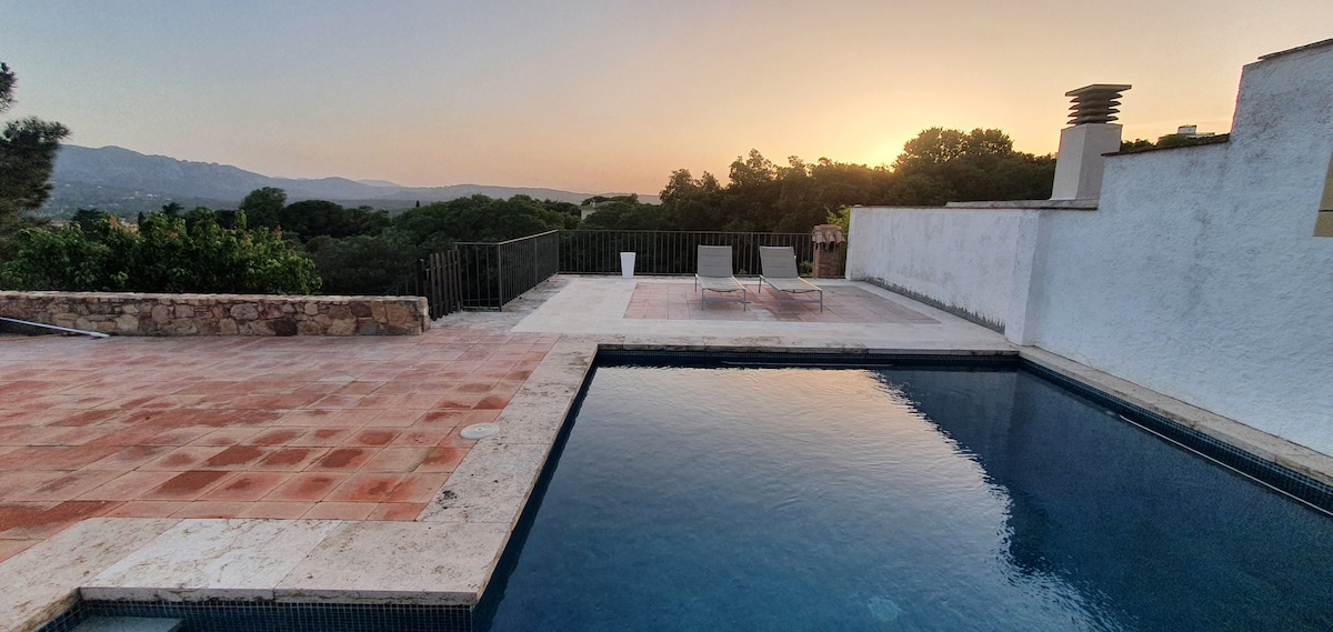 An outdoor space features a tranquil pool reflecting the sunset, surrounded by a tiled patio. Two lounge chairs are positioned near the water's edge, with a scenic view of the mountains in the background. The area offers a peaceful setting for relaxation.