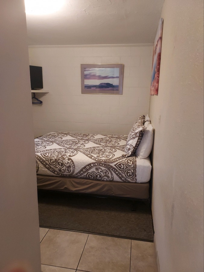A compact bedroom is shown with a decorated bed covered by a patterned quilt. A framed picture is hung on the wall above the bed, while a television is mounted on the opposite wall. Light-colored tiles cover the floor, enhancing the room's brightness.