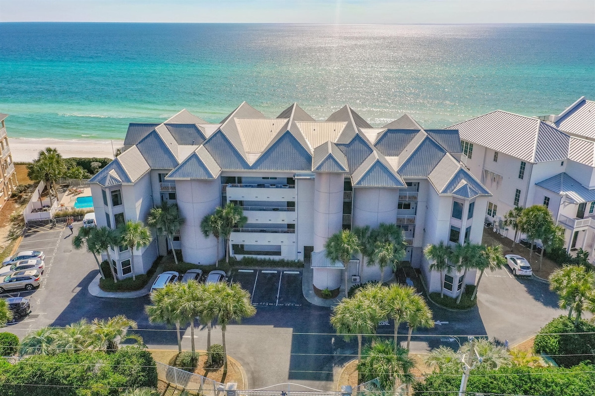 The exterior of a coastal condo building is displayed, showcasing a unique architectural design with a multi-angled roof. Lush palm trees line the property, and the vibrant blue ocean and sandy beach are visible in the background, offering a sense of tranquility.