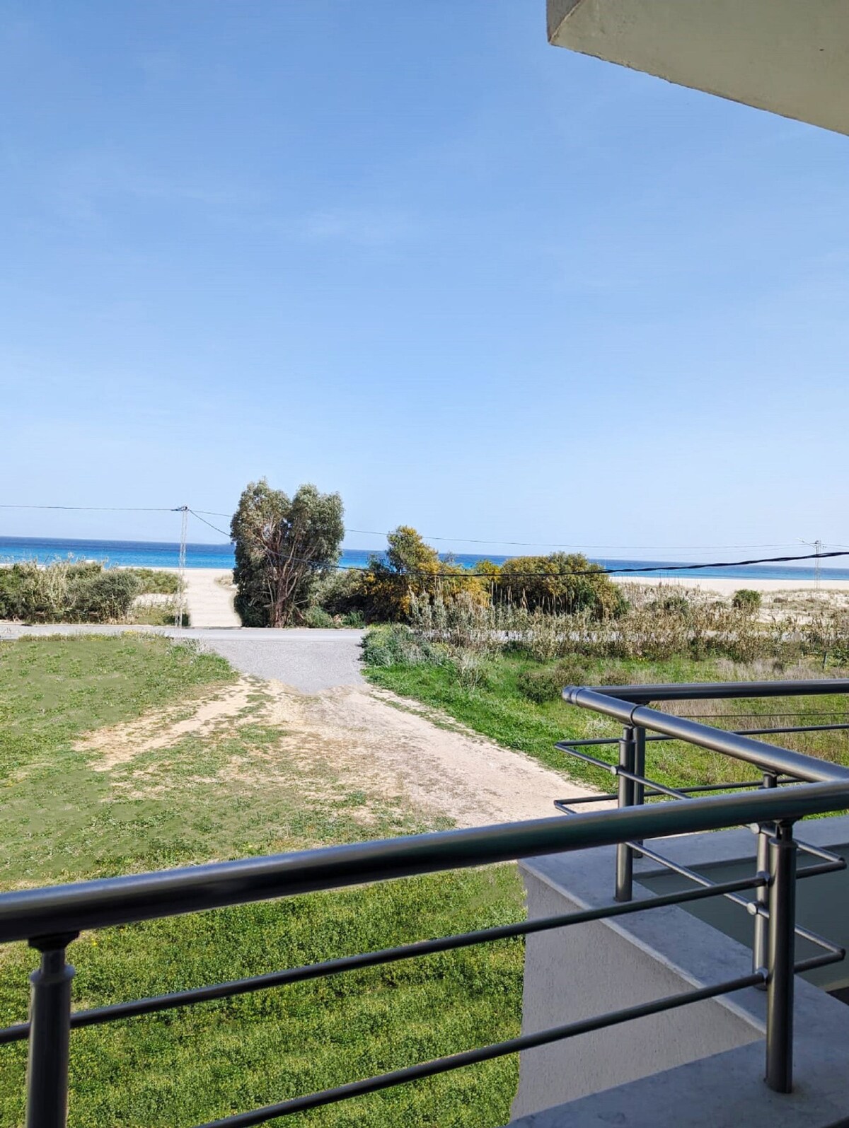 A view of the sandy beach is captured from the apartment's balcony. Lush greenery and walking paths lead towards the shoreline, while the calm blue sea stretches into the horizon under a clear sky.