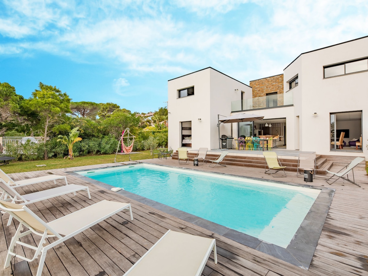 A modern villa exterior is highlighted by a rectangular swimming pool surrounded by wooden decking. Several lounge chairs are positioned for sunbathing, while lush greenery frames the property. A shaded terrace features seating for outdoor relaxation, enhancing the inviting atmosphere.