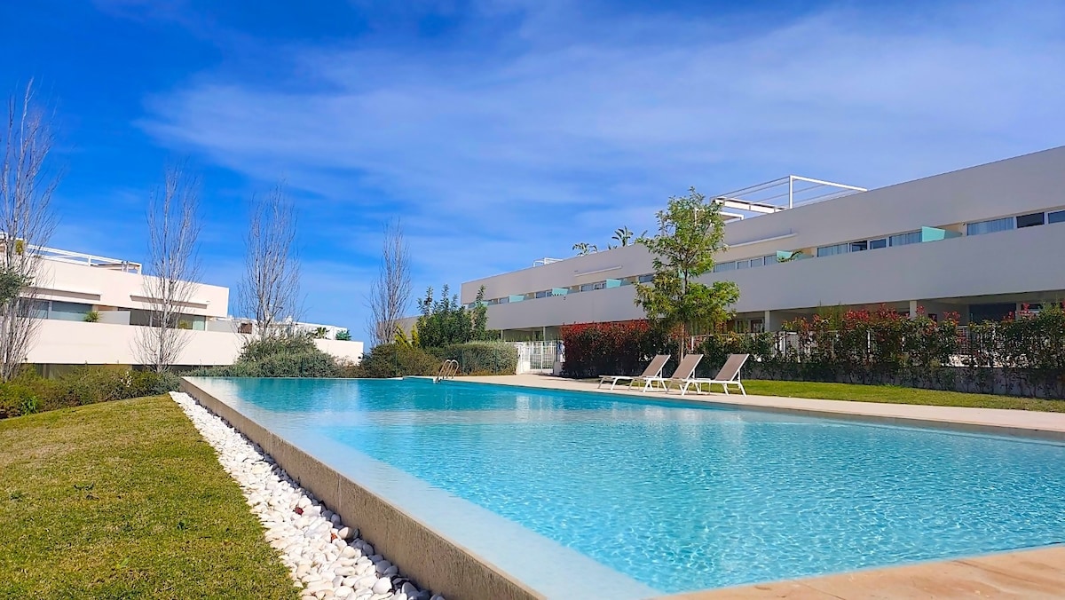 A serene outdoor pool reflects the clear blue sky, surrounded by well-maintained grass and decorative stones. Sun loungers are arranged elegantly nearby, enhancing the leisure space. Lush greenery and shrubs provide a sense of privacy, creating an inviting atmosphere for relaxation.