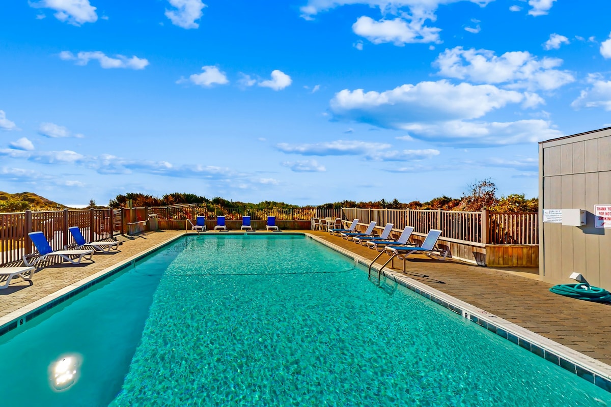 A turquoise pool is surrounded by a spacious deck furnished with lounge chairs, inviting relaxation. A wooden fence provides privacy, while distant greenery and a bright blue sky stretch out in the background, adding to the tranquil setting.