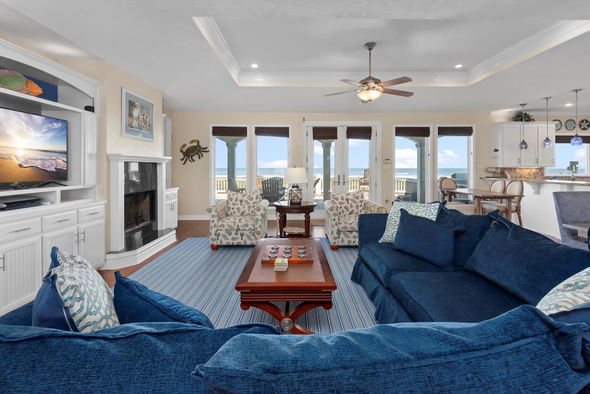 A spacious living area features a large navy sectional couch and patterned armchairs, providing ample seating. A flat-screen TV sits on a white cabinet, while large glass doors reveal panoramic views of the coastline. Natural light brightens the room, creating a welcoming atmosphere.