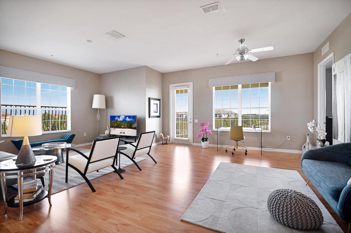 A spacious living area features large windows allowing natural light to fill the room. Stylish seating options include modern chairs and a comfortable sofa. A television is positioned on a sleek stand, with decorative elements like a floral arrangement and a textured area rug enhancing the space.