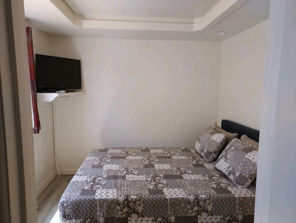 A compact bedroom is presented with a bed featuring a patchwork quilt and several pillows. A wall-mounted television is positioned above a small shelf, while natural light enters through a nearby window adorned with a patterned curtain.