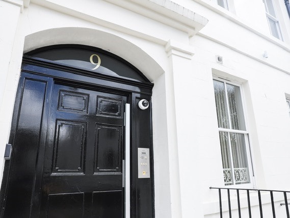 The entrance features a polished black door with large panels, marked with the number 9. A modern intercom is installed on the side, and a window with white framing partially reveals the interior. The surrounding white exterior walls highlight the door's elegance.