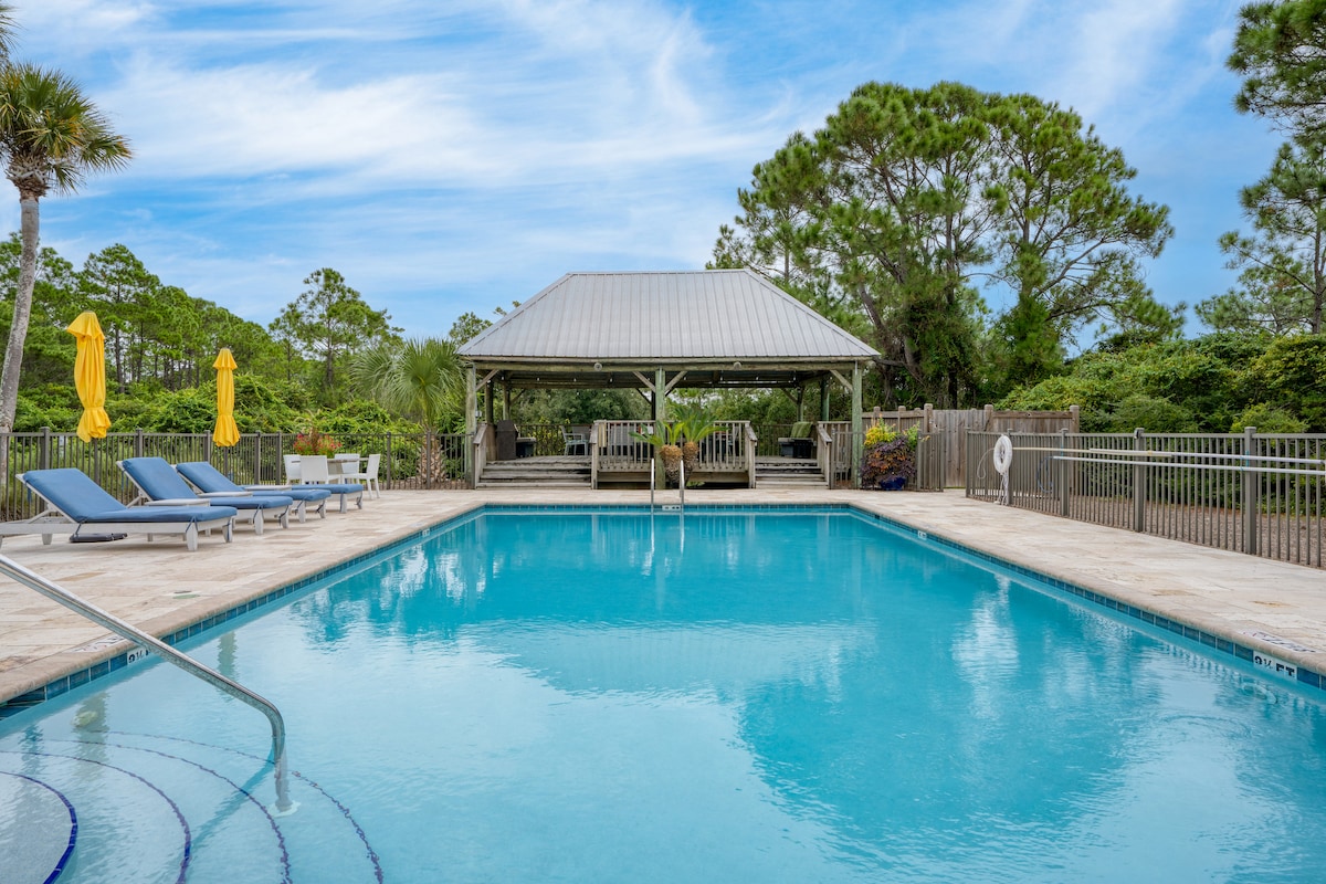 A community pool area features a clear blue pool bordered by lounge chairs under the sun. A spacious shaded cabana with seating is visible in the background, alongside lush greenery that enhances the serene atmosphere.