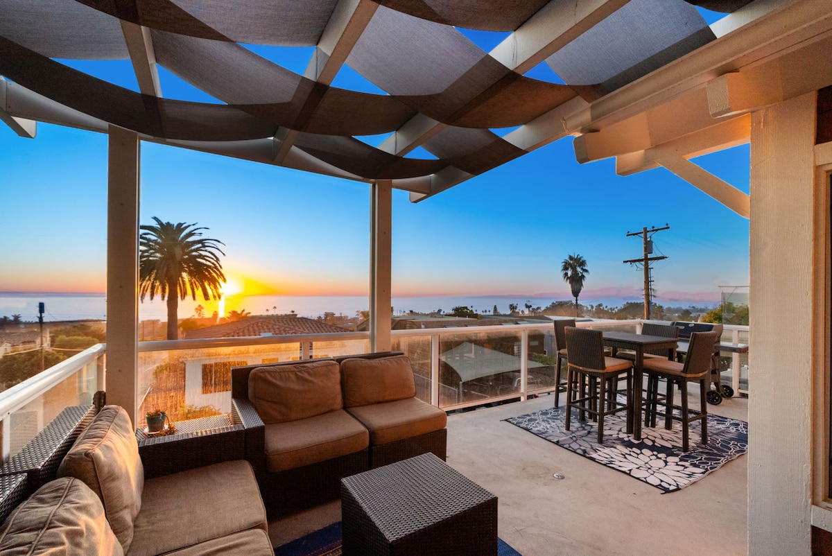 An outdoor patio is featured with a comfortable seating area, showcasing a sectional sofa and table. The space is shaded by a unique overhead design, with a stunning view of the ocean and sunset in the background, highlighted by palm trees.