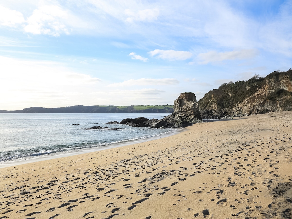 A sandy beach is depicted with gentle waves lapping at the shore. The coastline features a rocky formation on the right, while the horizon provides a backdrop of rolling hills under a clear sky. Footprints are visible in the soft sand.
